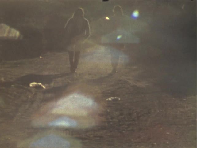 Two individuals walking on a dirt path in a hazy scene illuminated by light sources creating lens flares