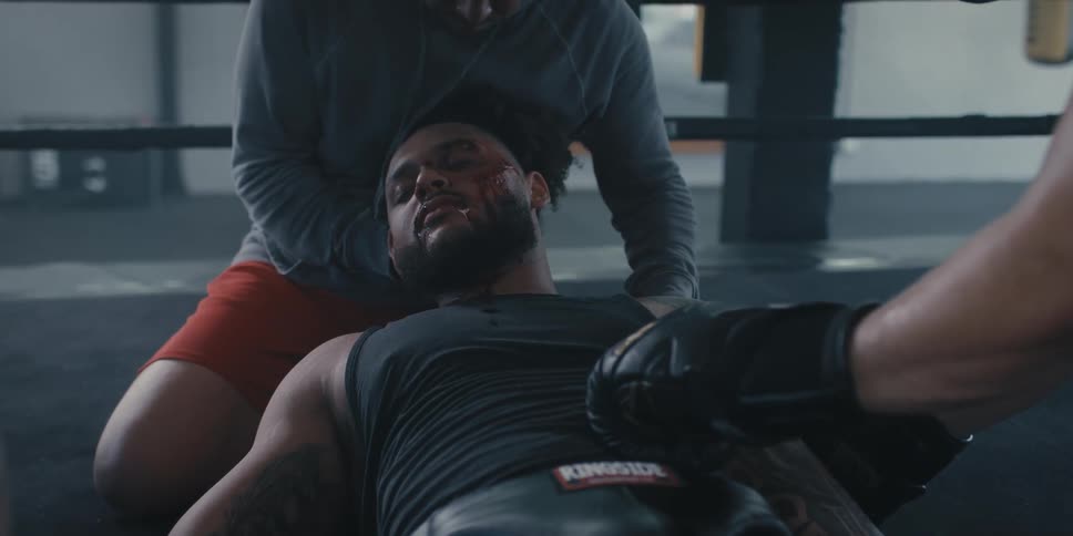 A boxer with bloodied wounds on his face lies on his back in the ring with another person kneeling beside him holding his head