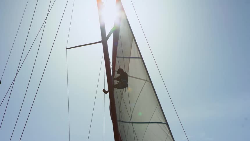 A person climbs the mast of a sailboat with the sun shining brightly from behind