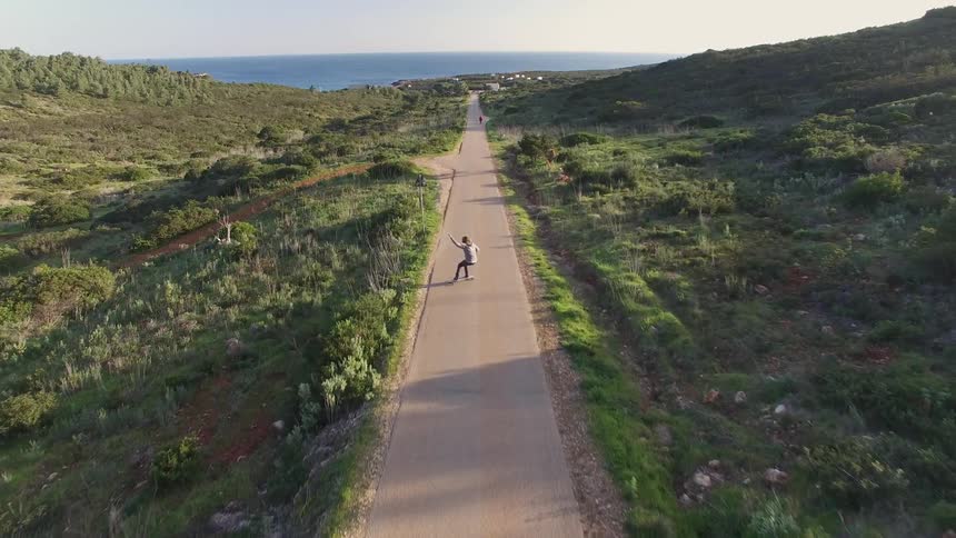 Aerial view of a person skateboarding down a narrow road surrounded by vegetation and with the sea in the background