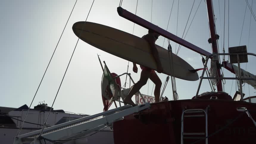 A young man carries a long surfboard on a sailboat