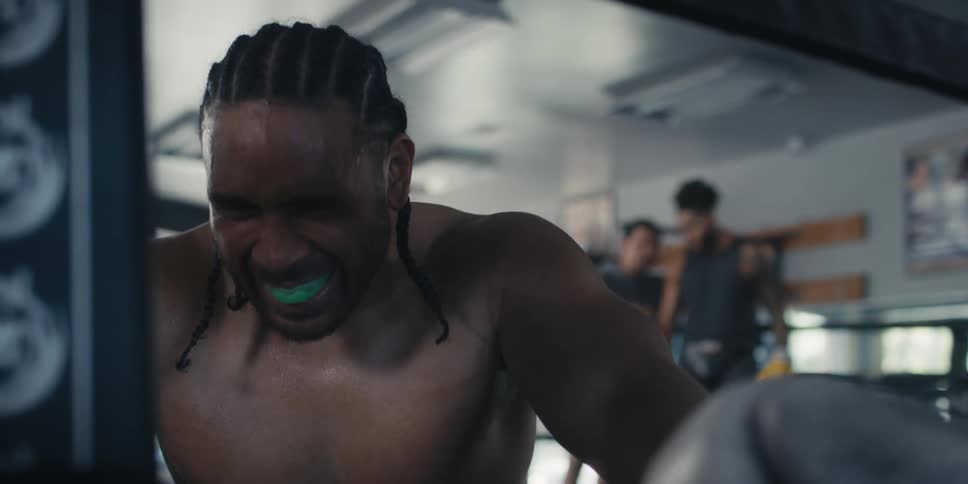 Man kneeling and taking a deep breath while standing in a boxing ring with another fighter in the background