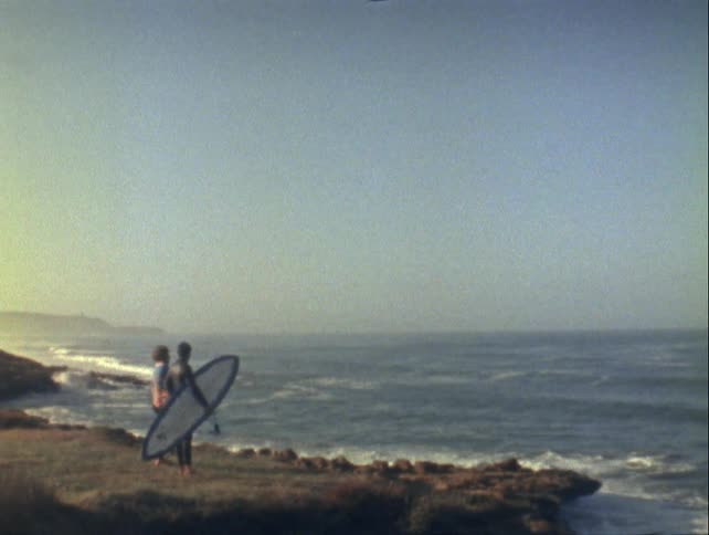 Two people stand on a rocky shore one of them holding a surfboard with the ocean in the background