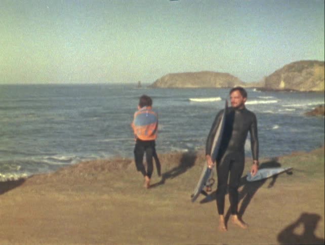 A pair of surfers on a sandy beach carrying surfboards with the ocean and a rocky coastline in the background