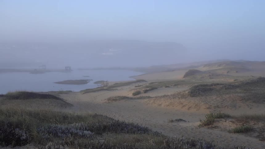 Sand dunes in the foreground with a body of water in the background and hills partially visible due to the fog