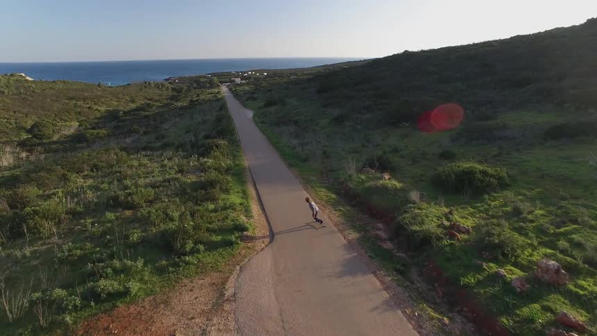 A person rides a skateboard on a paved road through a grassy landscape with the ocean in the distance