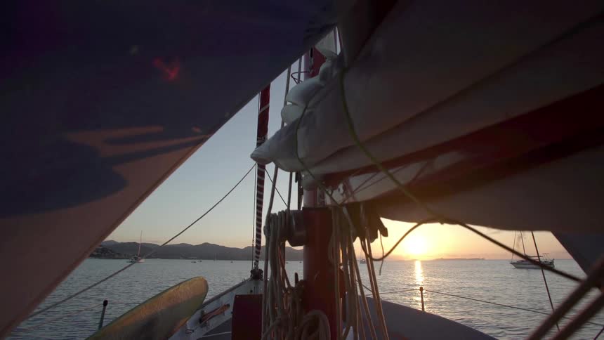 A sailboat deck at sunset with partially unfurled sails and calm waters