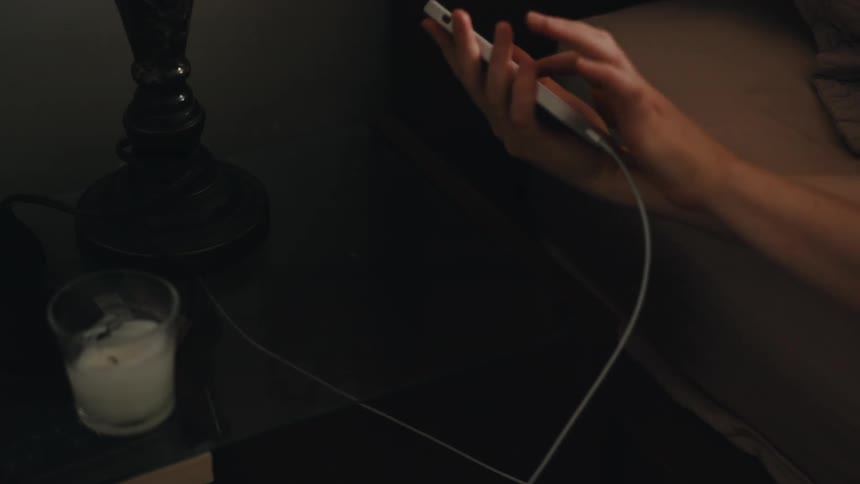 Closeup of a hand unplugging a smartphone from a charger next to the bed