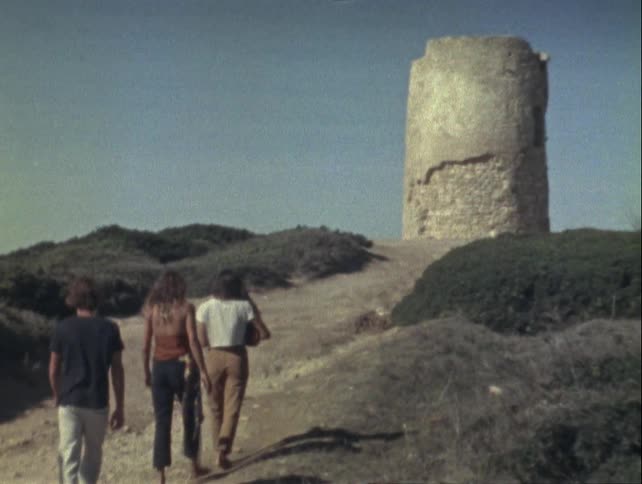 Four individuals walking towards an old stone tower on a hill with sparse vegetation under a clear sky