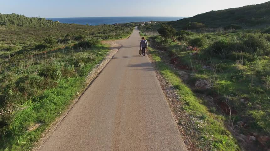 Man with a skateboard in his hand walks alone along a paved road surrounded by vegetation