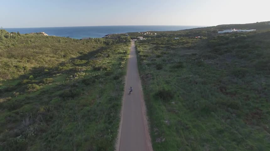 Aerial view of a person skateboarding on a road surrounded by vegetation and with the sea in the background