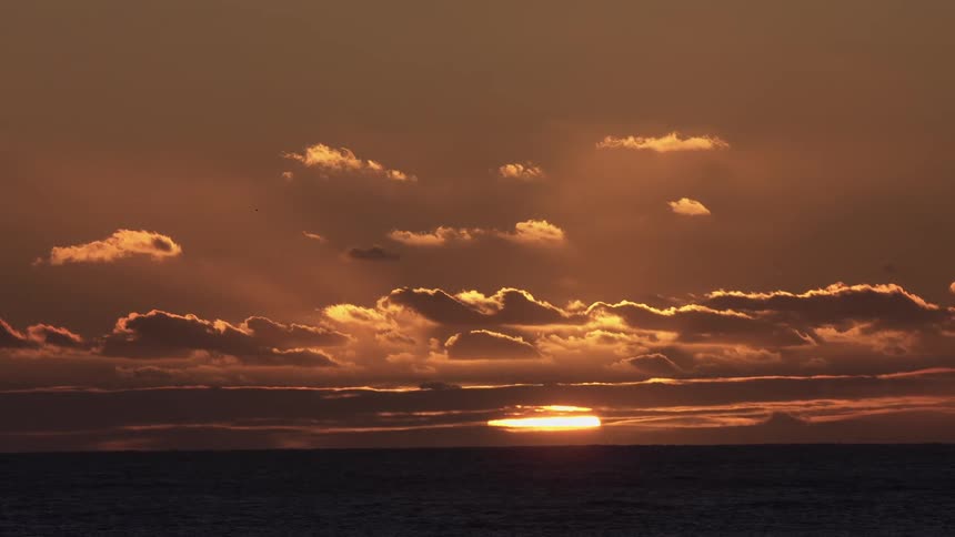 Sunset with the sun partially obscured by clouds near the horizon over a calm sea