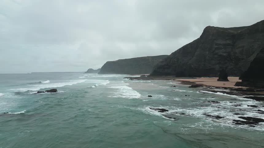 A wide shot of an ocean with waves crashing along a rocky sandy beach below dark cliffs on a cloudy day