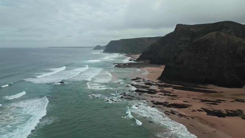 An aerial view of a rugged coastline with cliffs a sandy beach and waves breaking on the shore under an overcast sky