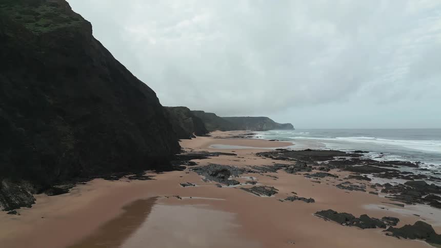 A coastal scene with a sandy beach strewn with rocks and seaweed flanked by tall cliffs under an overcast sky
