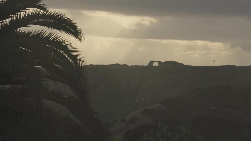 A natural rock arch is visible in the distance on a rugged hillside with rays of sunlight breaking through a cloudy sky and a large palm tree silhouetted in the foreground