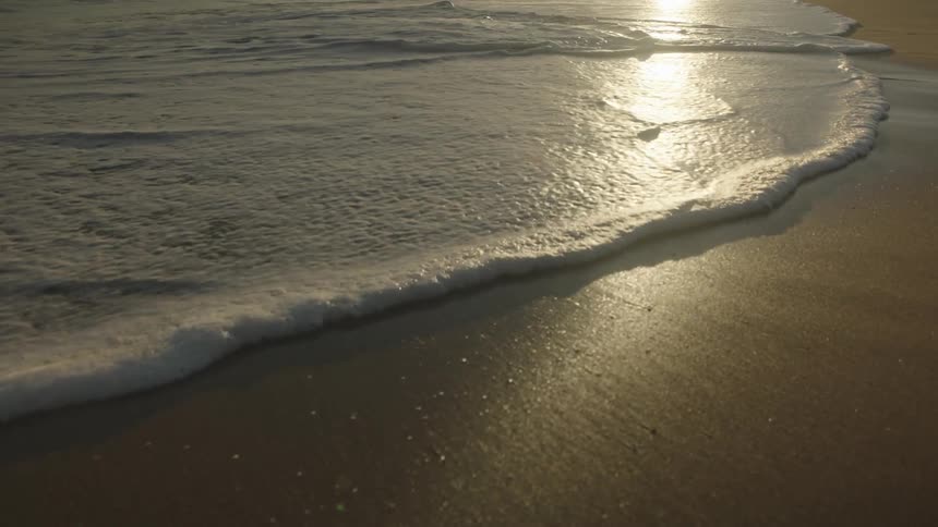 A closeup view of a beach where gentle waves are lapping onto the sand reflecting the sunlight