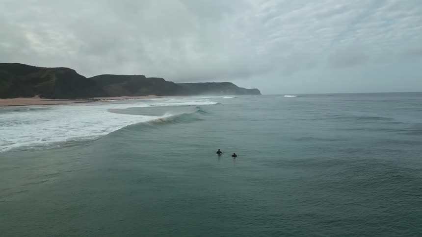 Two people on surfboards are in the water near a coastline with cliffs and cloudy skies in the distance