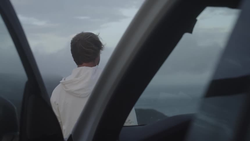 A man in a white hoodie stands outside of a car and stares off into the distance