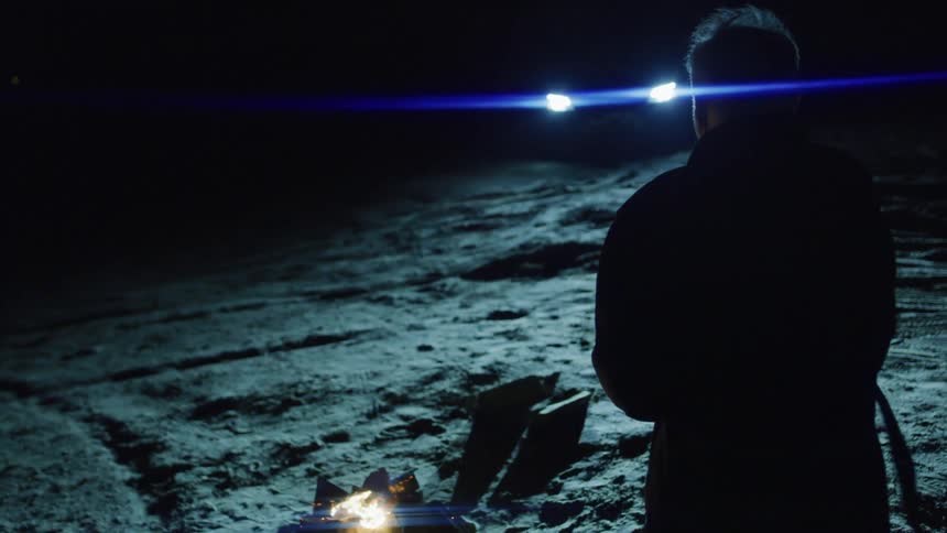 Man in front of a campfire at night illuminated by headlights of a car in the background
