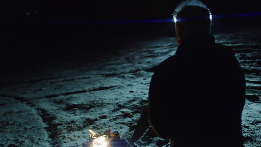 A person from behind looking at a small fire on the ground at night illuminated by a cars headlights