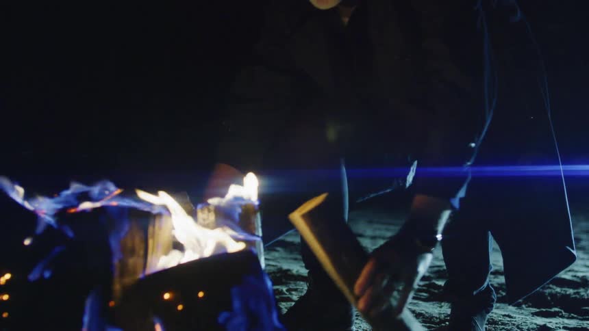 An elderly man carries logs to a bonfire burning on a beach