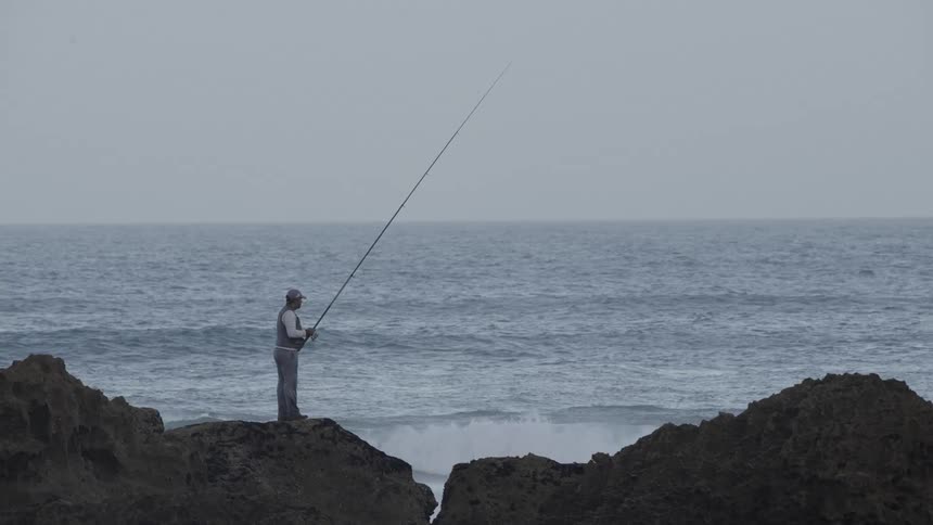 Man fishes while standing on the rocky shore on an overcast day