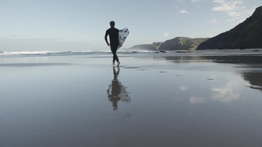 A male surfer in a wetsuit runs across a beach towards waves coming in from the sea