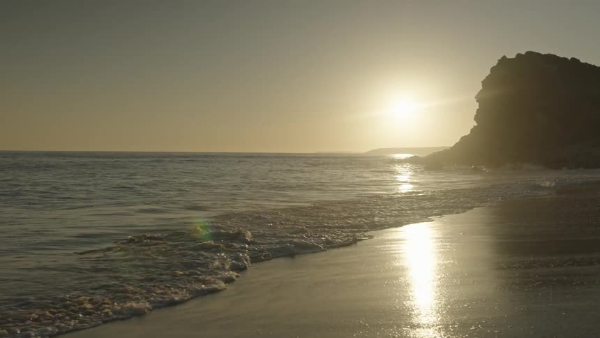 Waves roll into shore and over the beach near a promontory as the sun shines on the horizon in a hazy sky