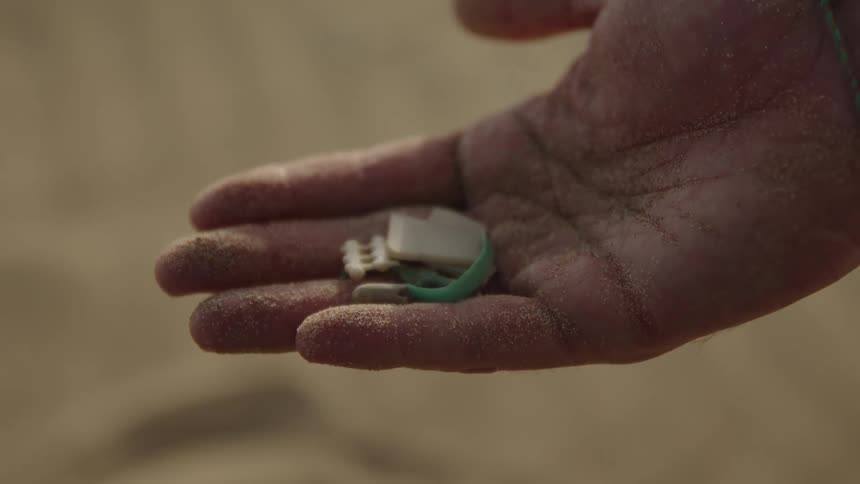 A hand covered in sand holds small pieces of plastic and debris with a green bracelet wrapped around the wrist