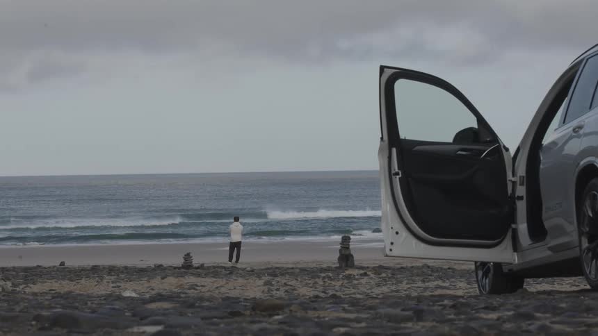 A car with its door open on a beach with a person standing near the waters edge
