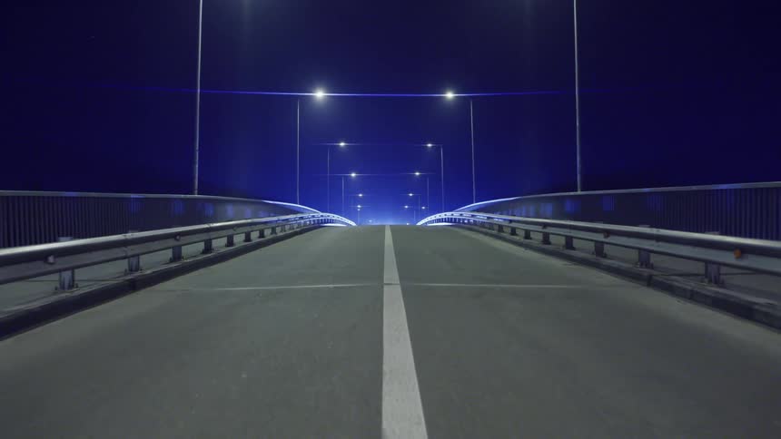A night scene of a vehicle with bright headlights on an empty bridge illuminated by streetlights