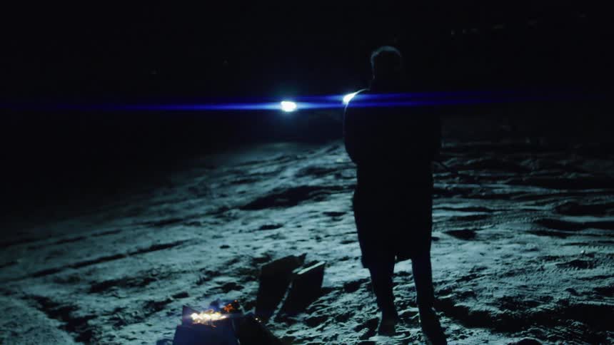 Silhouette of a man next to a campfire walking towards the glowing headlights of a vehicle in the background