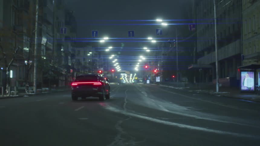 A car with red taillights driving on a wet city street at night with traffic lights ahead