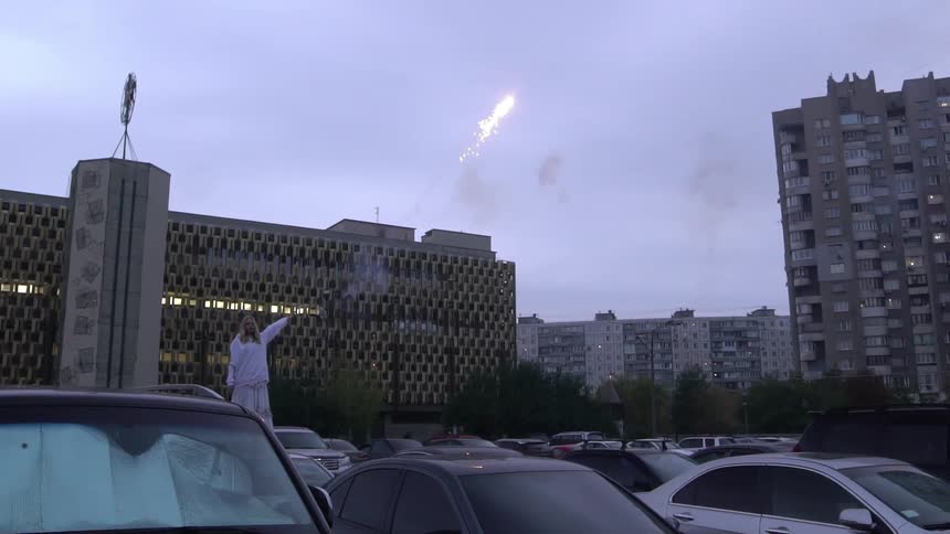 Woman shoots a roman candle up in a crowded parking lot outside a few buildings