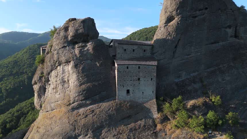 A stone building atop a tall rock in the mountains with green vegetation and rocky terrain around