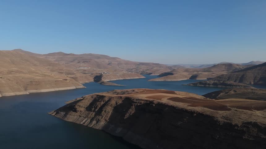 Deep blue body of water is surrounded by dry rolling hills and rugged terrain under a clear blue sky with distant mountains framing the background