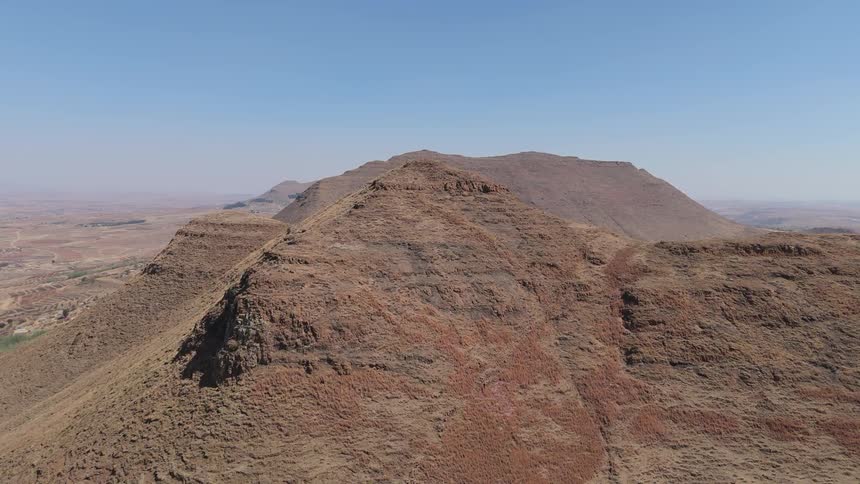 Rugged reddishbrown ridges in a dry mountainous landscape with expansive plains and distant peaks under a bright cloudless blue sky