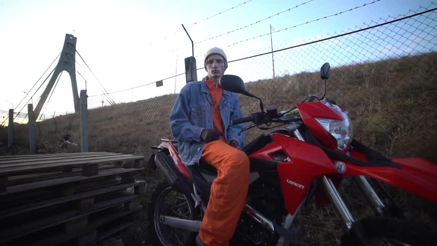 An individual in orange and blue attire sits on a red and white dirt bike next to a wooden pallet
