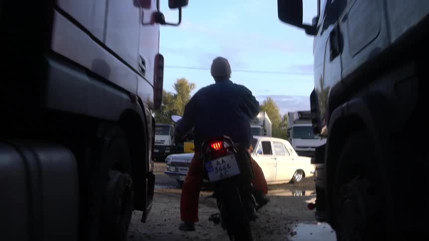 A man drives a motorcycle between trucks towards a woman reclined on a white car