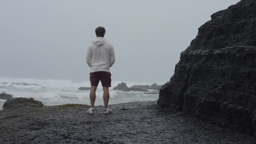 A man stands on a rocky coastline overlooking the ocean on a cloudy windy day