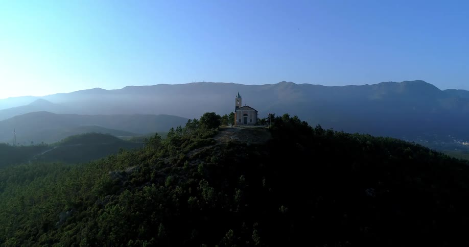 A church sits high on a forested mountain with a rural community sitting in a valley nearby