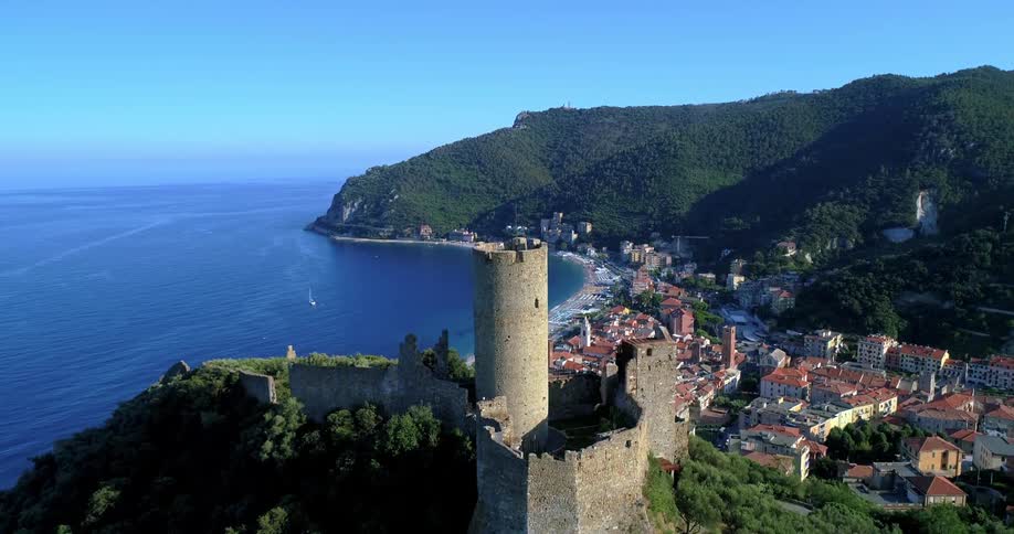 A stone castle stands atop a green hill overlooking a coastal town and the blue ocean