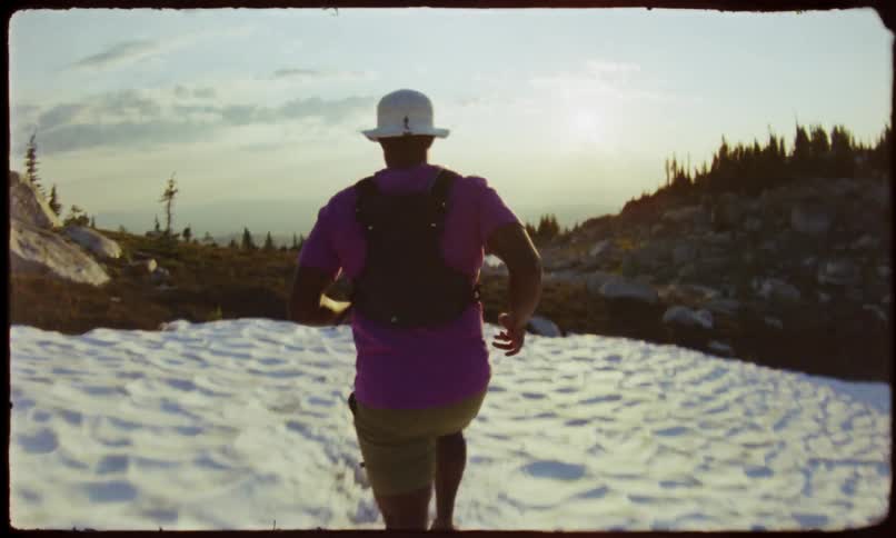 Man from behind wearing a purple tshirt running on a snowcovered terrain with trees and a sunset sky in the background