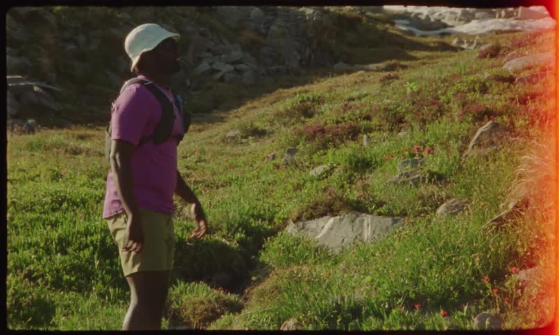 A man with a white hat and a pink shirt is running in an area with grass and lots of rocks and he stops and continues running after