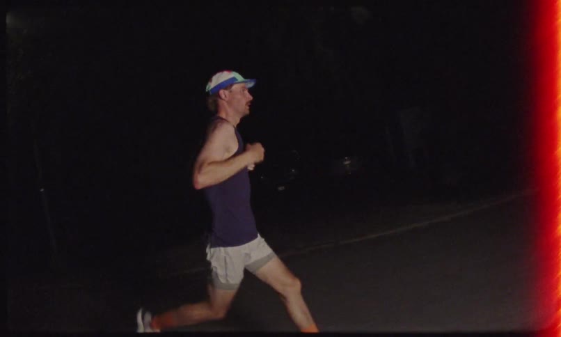 A man in a cap and black tank top is running on a street at night and staring straight ahead