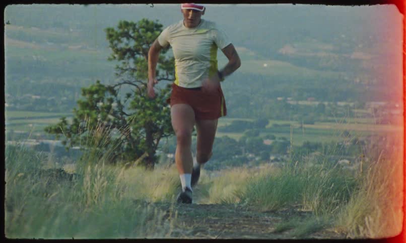 A woman is wearing a cap and shorts running forward in an area with grass and mountains in the background