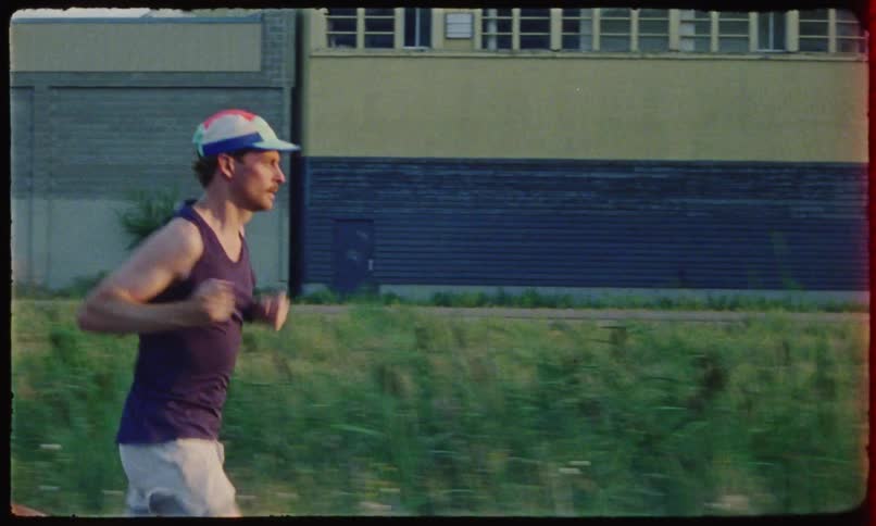 A white male wearing shorts and a hat jogs by a large warehousemanufacturing facility