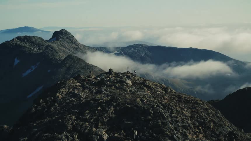 Group of people on top of a rocky mountain above the clouds with other mountains and a clear sky in the background