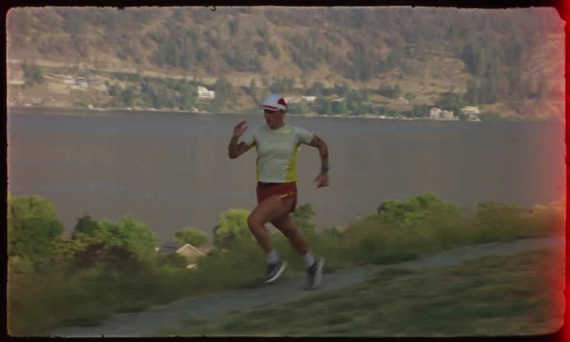 An woman is running along a path with a body of water and hills in the background captured on vintage film